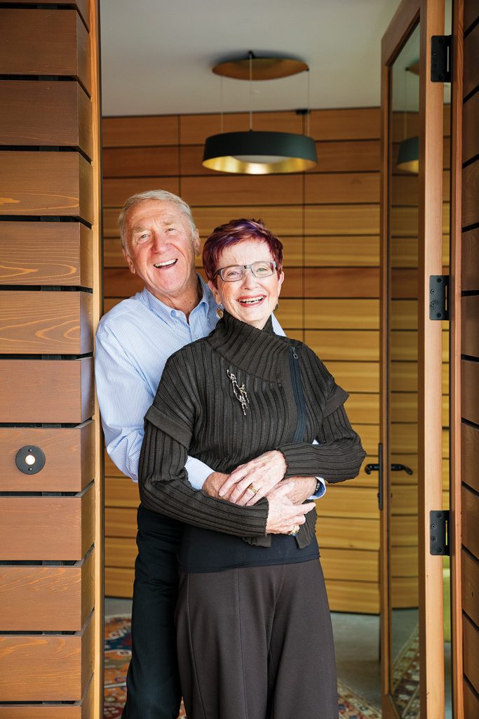 Norm and Margaret Pirk in their front doorway. Above right, the master bedroom has a neutral colour palette, with vertical and horizontal windows that bring in light and views of trees and sky.