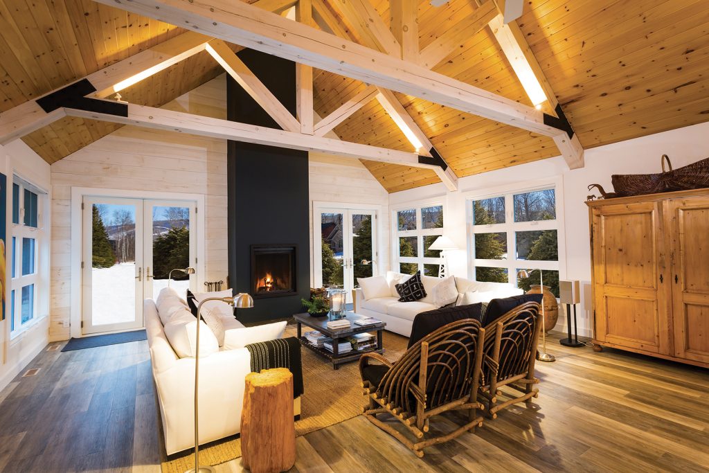 The ceiling in the open-concept living room is natural pine with a clearcoat, held up by king post trusses in whitewashed pine. The end wall is whitewashed pine, and floors are Armstrong vinyl with the look and texture of wood. Wood-burning fireplace by The Fireplace Stop, Schomberg, with metal surround by Thornbury Steel Fabricators finished in Farrow & Ball blue-black paint.
